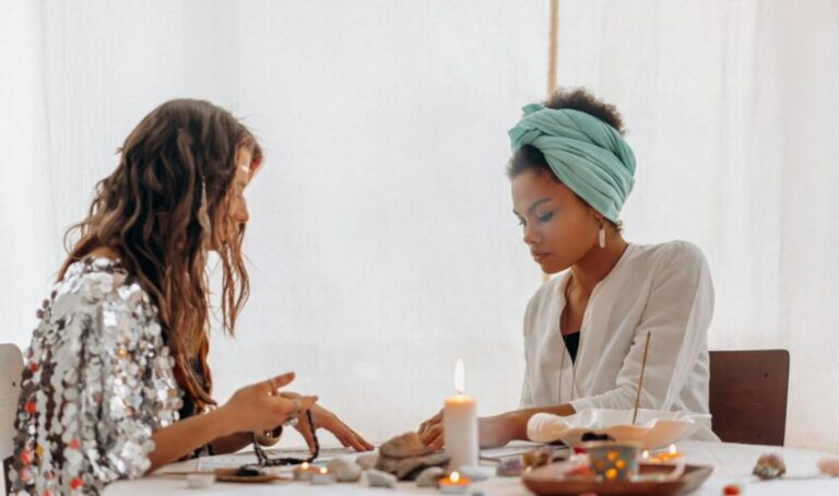 Two women participating in a spiritual ritual with candles and crystals on a table indoors.