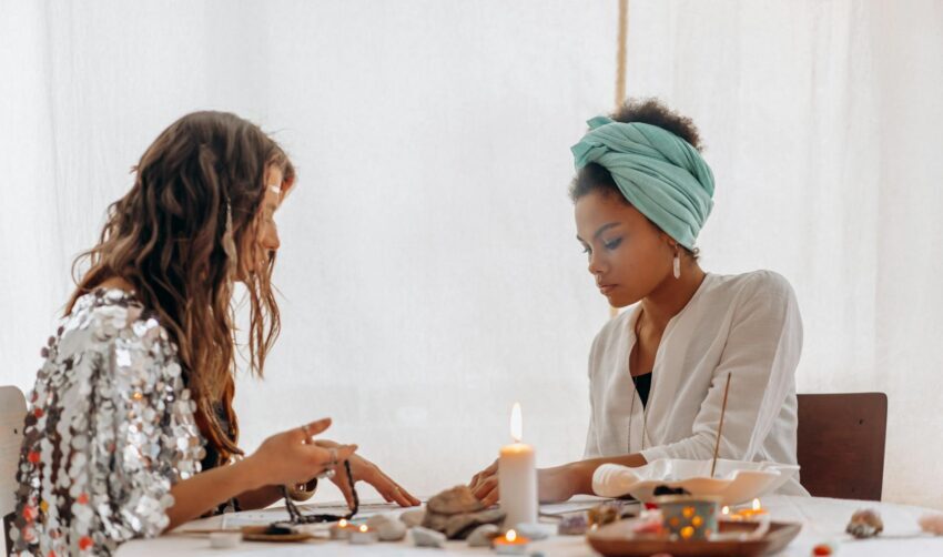 Two women participating in a spiritual ritual with candles and crystals on a table indoors.